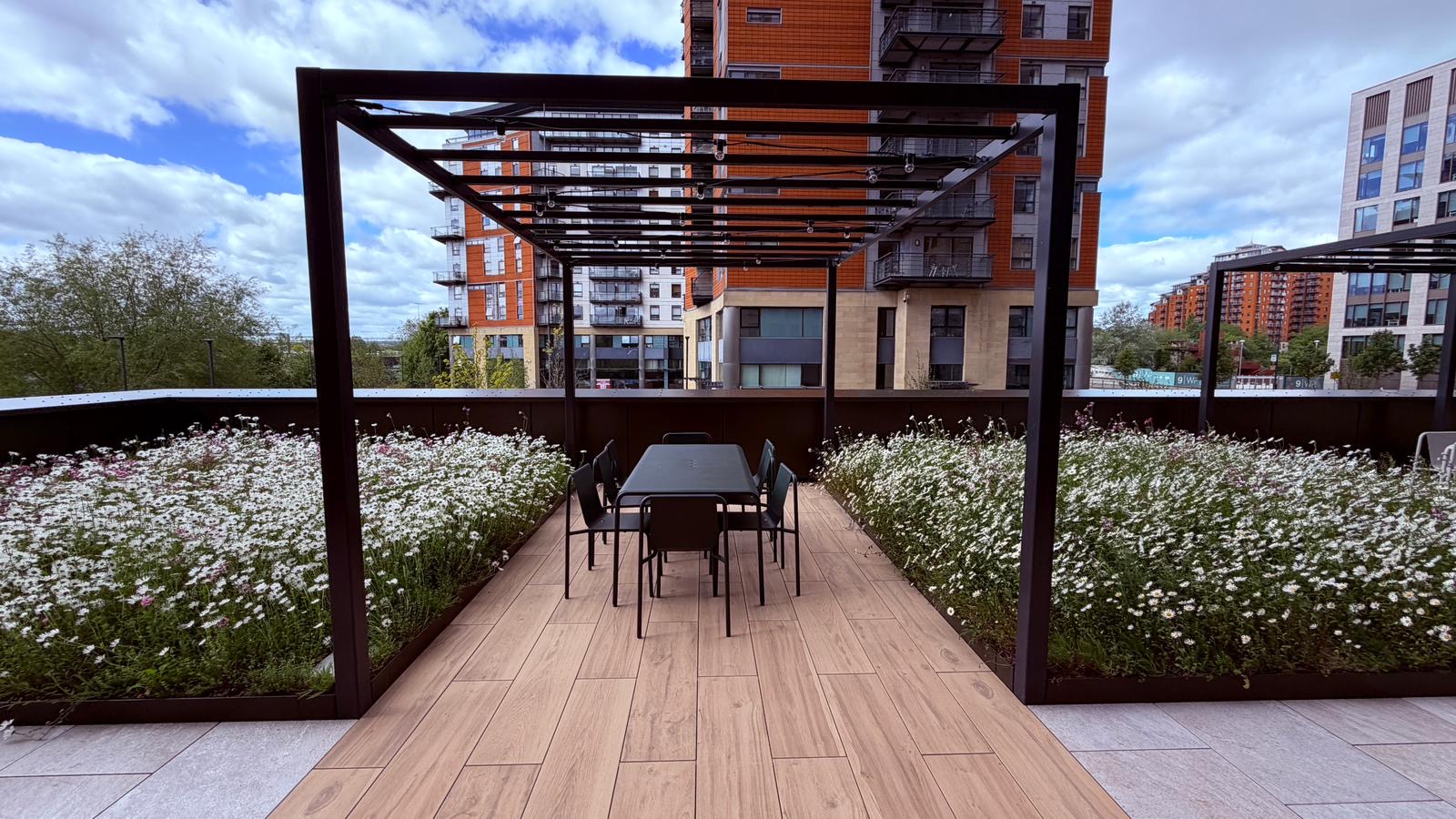 Steel pergola with dining table framed by white wildflower meadow planters on a podium terrace. — comparable to schemes we deliver in Hackney
