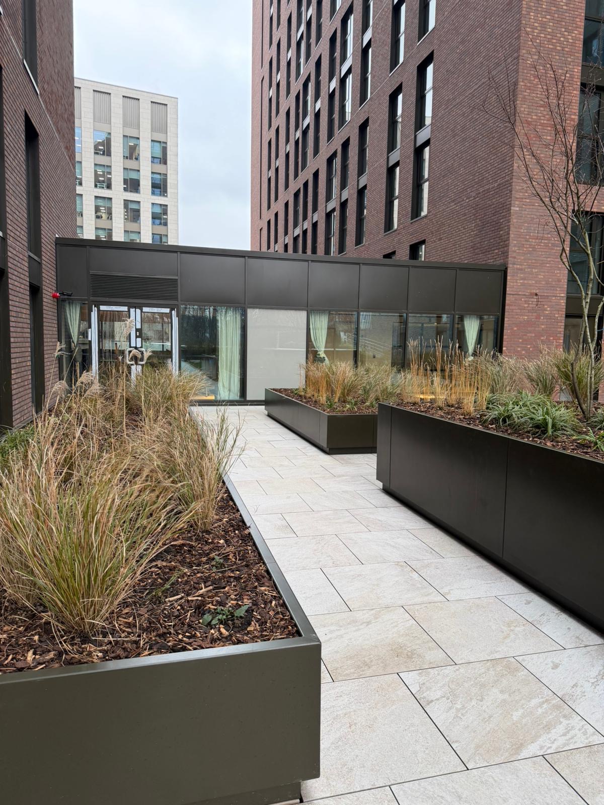 Matte black aluminium planter run lining a residential courtyard walkway with mixed grass planting. — comparable to schemes we deliver in Kensington & Chelsea