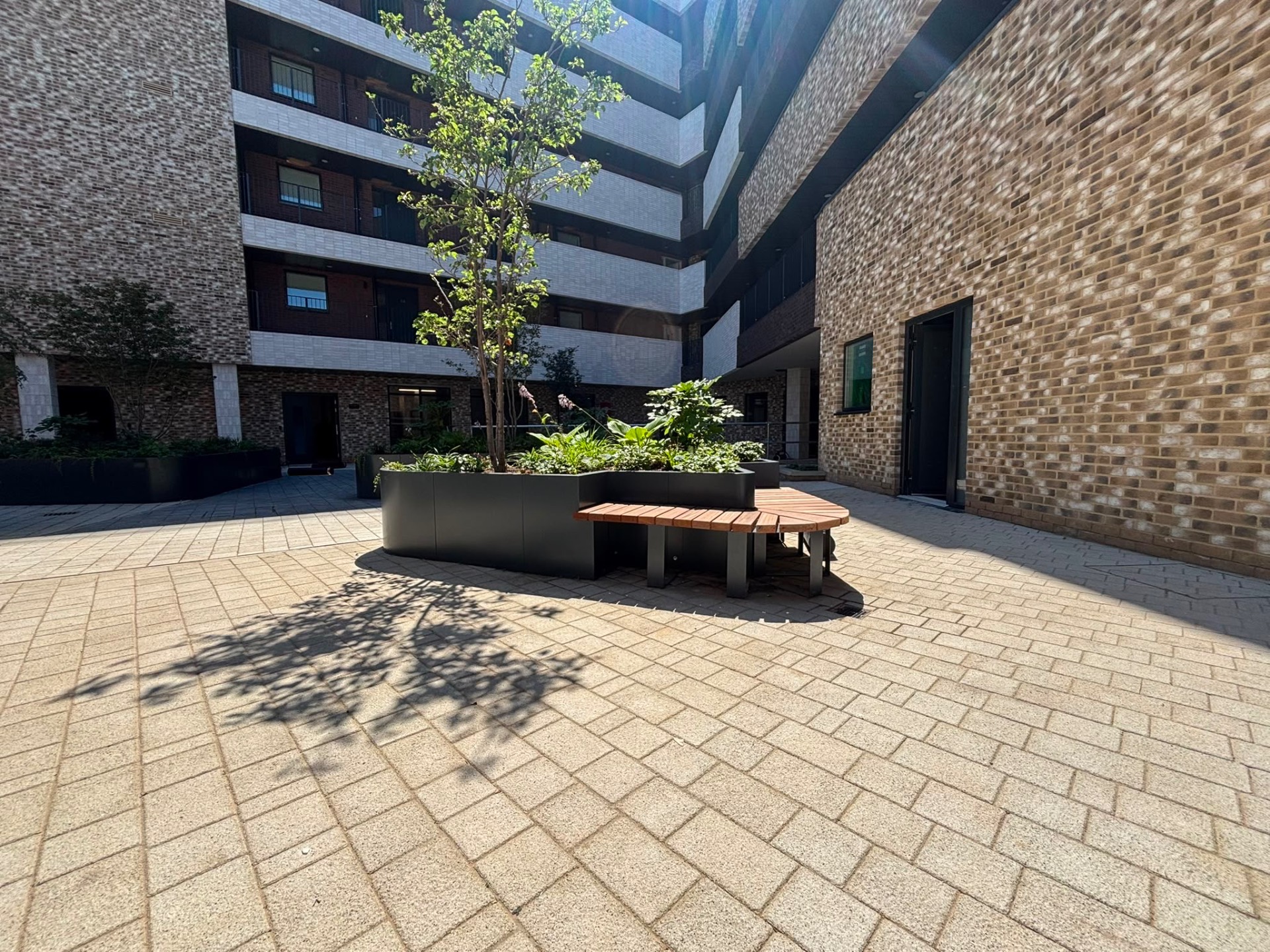 Angular matte black planter with integrated timber bench wrapping a multi-stem tree in a residential courtyard. — comparable to schemes we deliver in Tower Hamlets