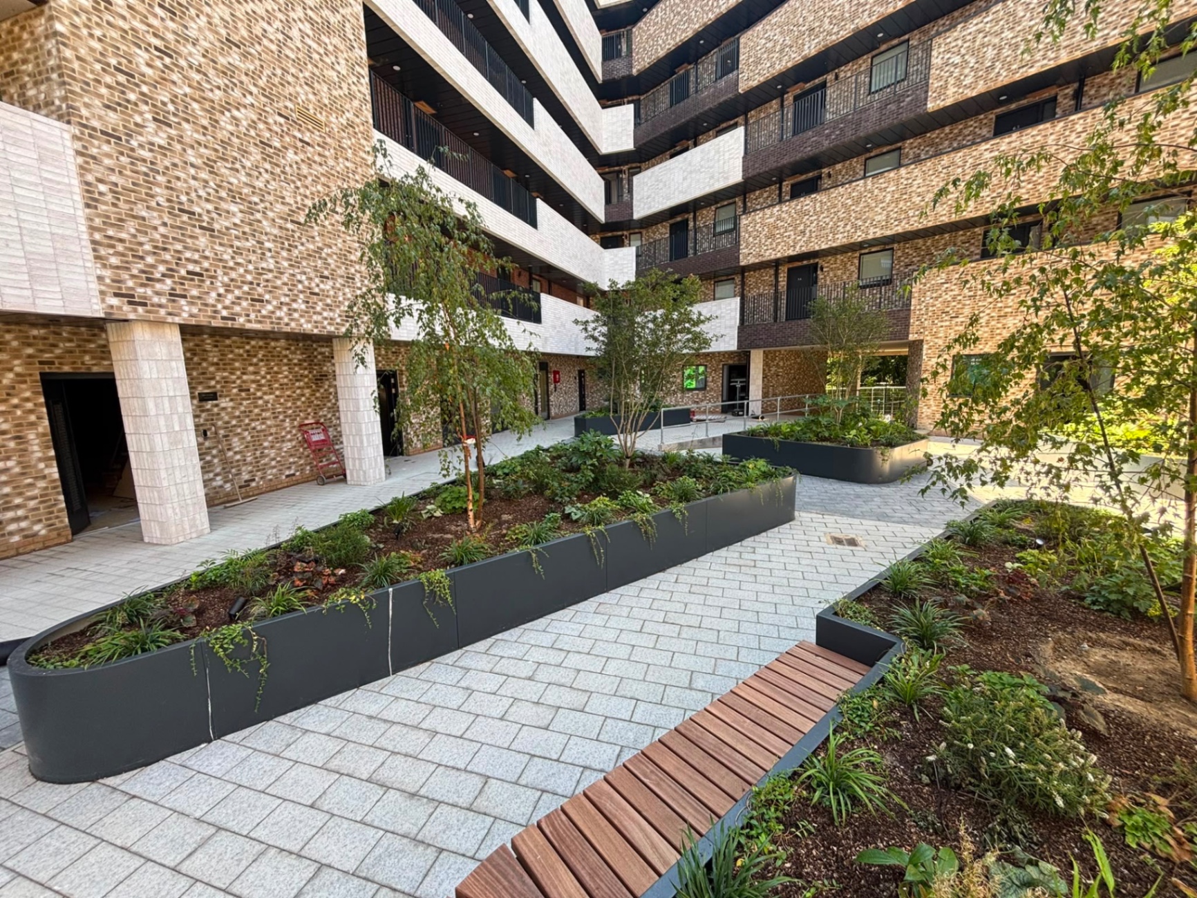 Bespoke metal planters of the kind we fabricate and install in Wandsworth — Serpentine matte black planter run with mixed shrub and grass planting winding through a new-build courtyard.